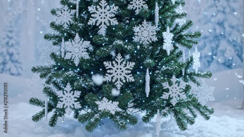 Festive Christmas tree adorned with large white snowflake ornaments and twinkling blue LED lights in a snowy winter forest landscape background.