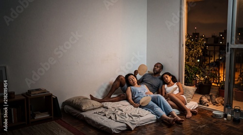 Cozy Evening Relaxation: A diverse group of people relaxes on a floor mattress in a cozy apartment, enjoying a peaceful evening with a city view outside the window.