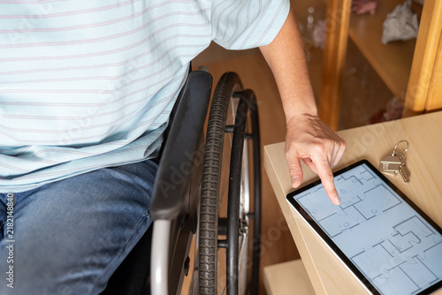 Person in wheelchair reviewing accessible home floor plan on tablet while holding house keys, planning independent, inclusive living with mobility adaptations and smart design features