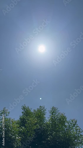 Static vertical shot of bright sun above lush green treetops under a clear blue sky.