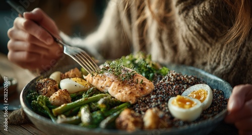 Close-up of a person holding a fork, preparing to eat a healthy meal featuring grilled salmon, quinoa, and assorted vegetables on a rustic wooden table