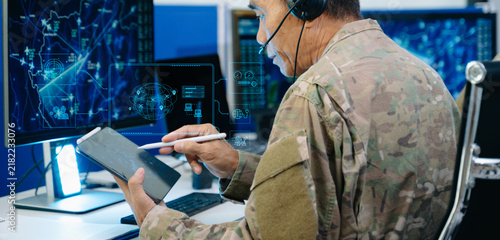 Senior military officer in camouflage using a tablet to analyze tactical radar data alongside his team in a modern cybersecurity command center.