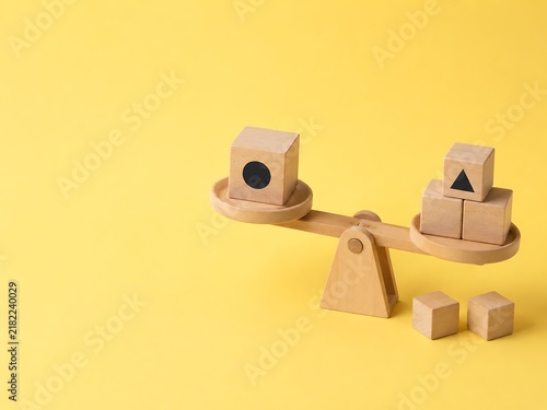 Wooden balance scale on a yellow background, with a block, featuring a circle, on one side, and multiple blocks, with triangles on, the other