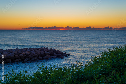 Golden sunrise over a calm blue sea with a rocky breakwater and green coastal vegetation, vivid orange sky glowing above dark clouds on the horizon, a peaceful and inspiring seascape in dawn light