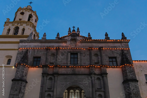 Low angle view of an old stone church facade and bell tower lined with strings of warm festive lights, glowing against a deep blue evening sky in Coyoacan, Mexico City.