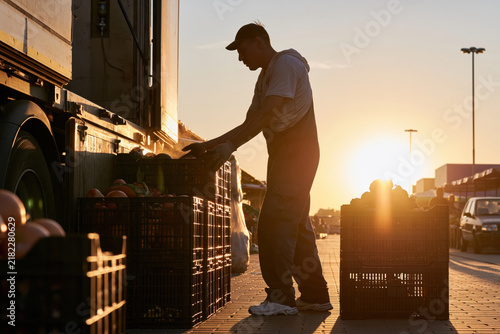 Farmer unloading crates of vegetables from truck at sunrise on market street