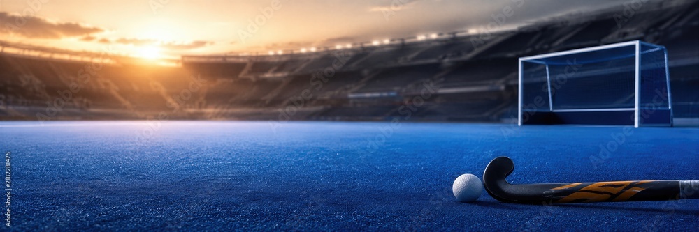 Fototapeta premium Field hockey stick and ball on blue pitch near goal at sunset. Empty stadium with dramatic sky at world field hockey championship. Sport equipment, tournament and competition. Banner with copy space