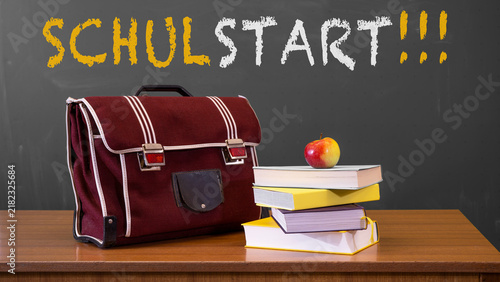 Schulstart, Schulbeginn, Einschulung Grußkarte, Karte mit deutschem Text - Schulranzen und Bücher auf Lehrerpult mit Schultafel im Hintergrund