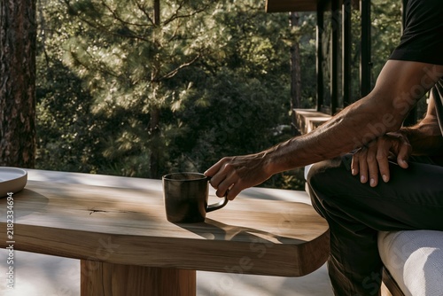 Closeup of a man's hand placing a stoneware coffee mug on a chunky teak table in a forest retreat with dappled light filtering through trees
