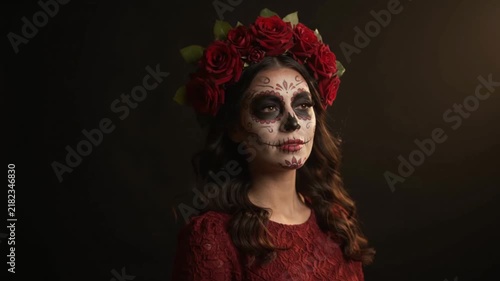A woman wearing elaborate sugar skull face paint and a crown of red roses looks into the distance, creating a dramatic cinematic scene inspired by Mexican tradition.