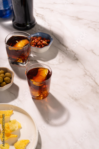 Fotografie Vermouth glasses with soda and a twist of orange on a marble table with snacks like olives, peanuts and potato chips