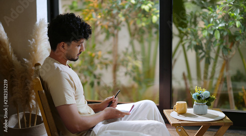 Indian calm young adult gen z man sitting on chair enjoy day resting indoor room. Male boy hold pen writing book new work notes plan ideas spend time take casual job break home house place