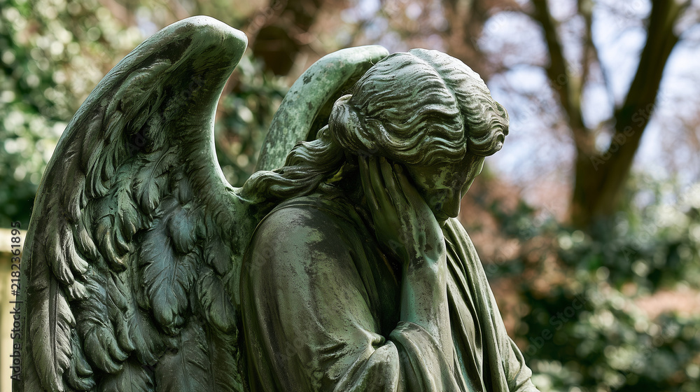 Fototapeta premium Side view of an antique angel sculpture bowing its head in grief, hand covering part of the face while detailed feathered wings and green weathered patina emphasize decades of outd