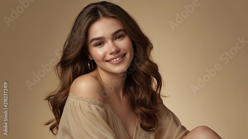 Young woman with wavy brunette hair smiles warmly in a neutral studio portrait setting.