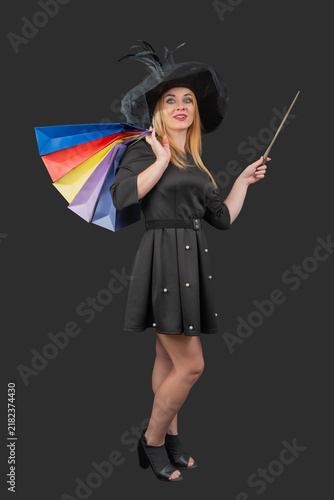 Obraz na plátně Smiling woman wearing a black witch hat and dress, carrying colorful shopping ba