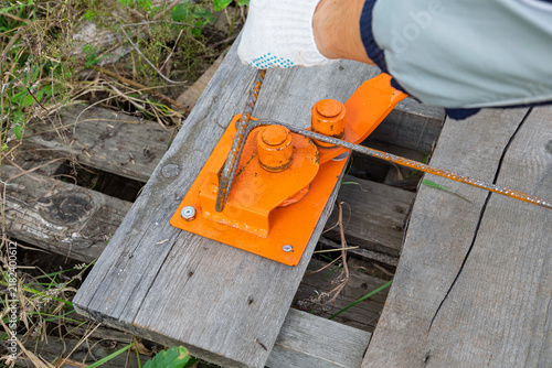 Fotografie Man worker using orange manual rebar bender to shape steel rod on wooden base at construction site