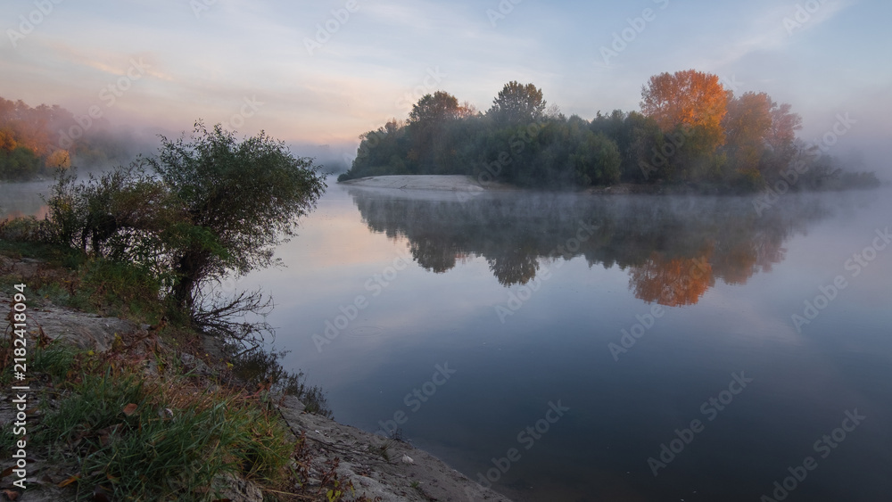 Obraz premium calm, misty river reflecting lush green and autumn-colored trees under a soft, hazy sky.