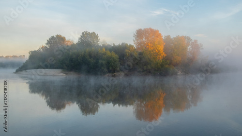 Obraz na plátně A misty morning landscape captures a small island with colorful autumn trees beautifully reflecting on the calm, mirror-like water surface