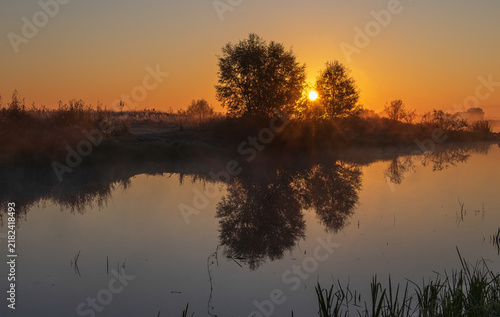 A beautiful sunrise casts a golden glow over a calm river, reflecting the silhouettes of trees on its peaceful water