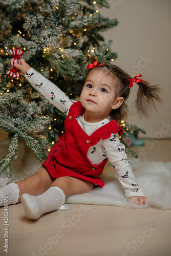 A young girl with brown hair and pigtails sits by a decorated Christmas tree. She wears a red dress and holds a small ornament, smiling happily.