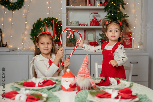 Two young girls celebrate Christmas at a festive table. One girl holds a candy cane, while the other stands beside her. The table is decorated with holiday items.