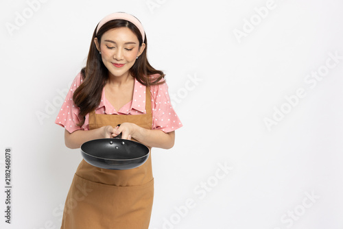 Happy young Asian woman wearing apron looking down at frying pan isolated on white background, housemaker, chef, restaurant cook, home cooking, food smell, recipe, and kitchenware concept