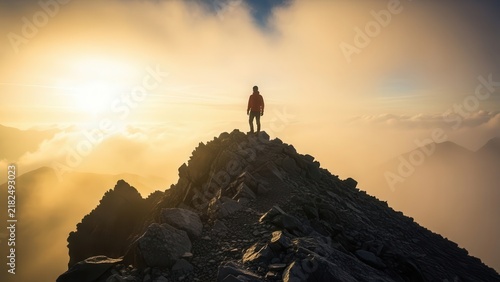 Person standing on a mountain ridge at sunrise. Golden light illuminates the clouds and distant peaks.