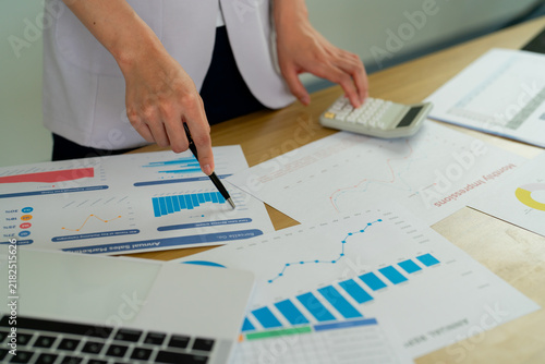 Business analyst pointing pen at growth chart on desk covered with financial reports, sales graphs and pie charts, analyzing annual data with calculator nearby