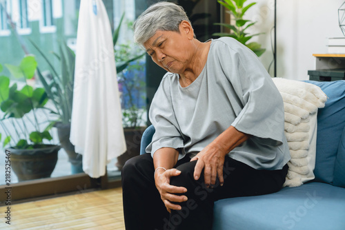 Elderly Asian woman sitting on a sofa at home, holding her painful knee, experiencing joint discomfort and mobility problems.