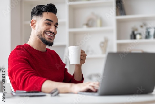 Cuadro en lienzo Smiling Young Middle Eastern Man Enjoying Coffee While Working On Laptop In Home Office, Focused On Task
