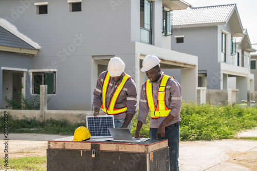 Two construction workers examine a solar panel and laptop outdoors at a residential housing development site, reviewing sustainable energy installation plans during daytime project management.