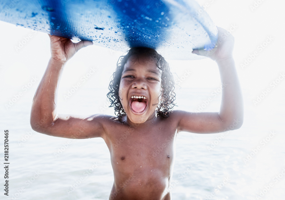 Obraz premium Joyful Black boy holding a blue surfboard overhead at the beach