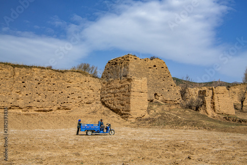 A farmer driving a small three-wheeled vehicle across dry farmland near eroded earthen structures in northern China. 