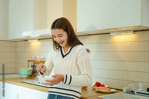 Fotografie Cheerful young Asian woman enjoys preparing a meal in a bright modern kitchen, i