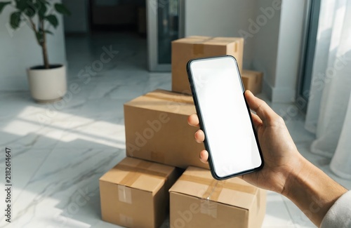 Hand holding smartphone with blank white screen near cardboard moving boxes in new home