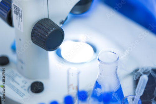 Photography Laboratory glassware filled with bright blue reagent arranged at a brightly lit laboratory workbench, surrounded by flasks of glowing blue reagent and racked glass test tubes