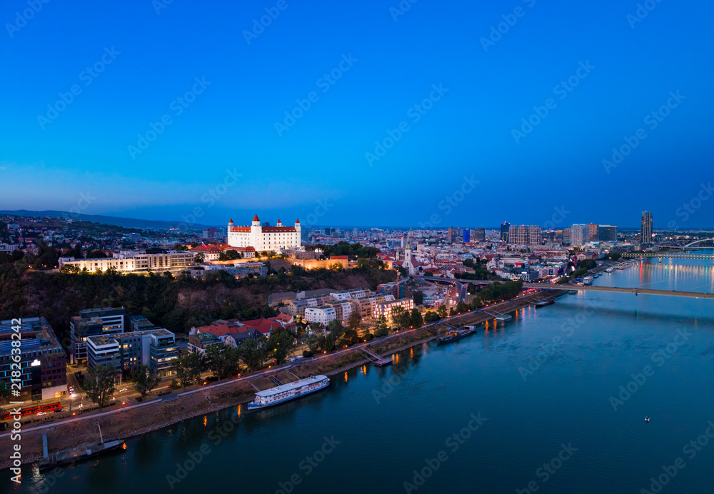 Obraz premium Bratislava skyline at dusk with Danube River and city lights visible. City lights shine along the Danube River in Bratislava during dusk. The castle is on the hill.
