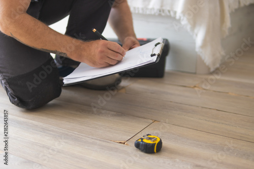 Contractor inspecting house floor damage, measuring cracks with tape measure and writing notes on clipboard, assessing property for repair and home renovation