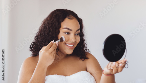Obraz na plátně Smiling Black Woman Applying Makeup With Brush In Front Of Round Mirror At Home, Enjoying Self-care Routine In Bright Interior Setting For Beauty And Personal Care Concept