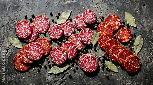 Slices of various cured sausages arranged on a dark stone background with bay leaves and peppercorns