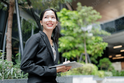 Smiling businesswoman holding a tablet with stylus in a modern green office courtyard, showing professional confidence, digital work, productivity, career success, leadership, and corporate lifestyle.
