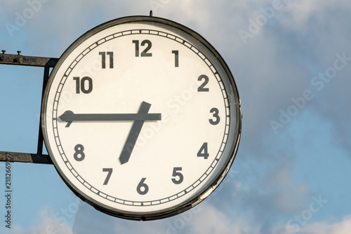 Round wall clock with black hour and minute hands showing 10:10 against a blue sky with clouds, featuring a classic design and clear numerals for easy readability