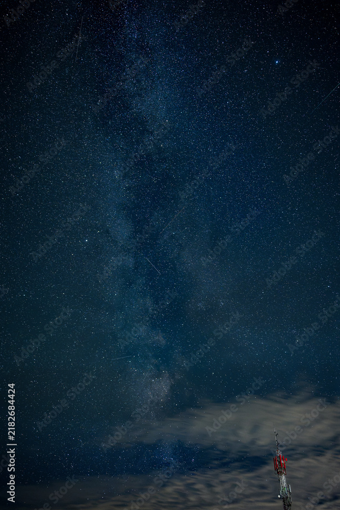 Fototapeta premium Milky Way stretching across a star-filled August night sky above a telecommunications tower and drifting clouds, combining astronomy, wireless technology and modern infrastructure.