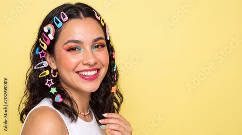 Happy young woman with colorful hair clips and bright makeup smiling at camera against yellow background with eyeshadow looks directly