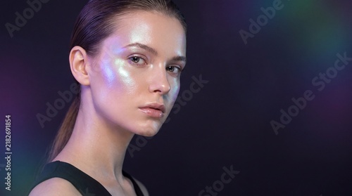 Young woman with holographic makeup posing against a dark background with colorful light flares with glowing holographic highlighter