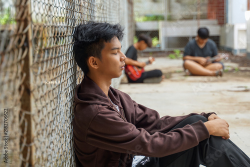 Young Asian male leaning back with eyes closed against a wire fence while friends sit blurred in the background outdoors.