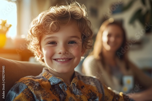Young boy with curly hair smiling brightly while sitting at home with his mother in the back