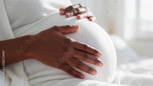 Close-up of a pregnant black woman gentle hands gently caressing her large round belly in white ribbed knit maternity clothing. Future motherhood and prenatal care concept