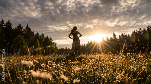 A graceful woman stands in a sunlit meadow surrounded by wildflowers, her silhouette glowing under golden rays. The warm light and dynamic sky evoke freedom, nature, and serenity.