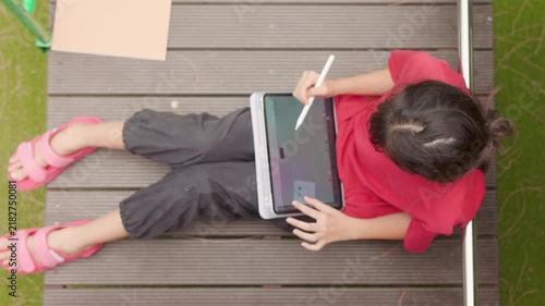 Top view of an Asian kid enjoying a relaxing hobby, drawing digital art on a tablet. Selective focus captures the elementary student spending casual leisure time at an outdoor school.